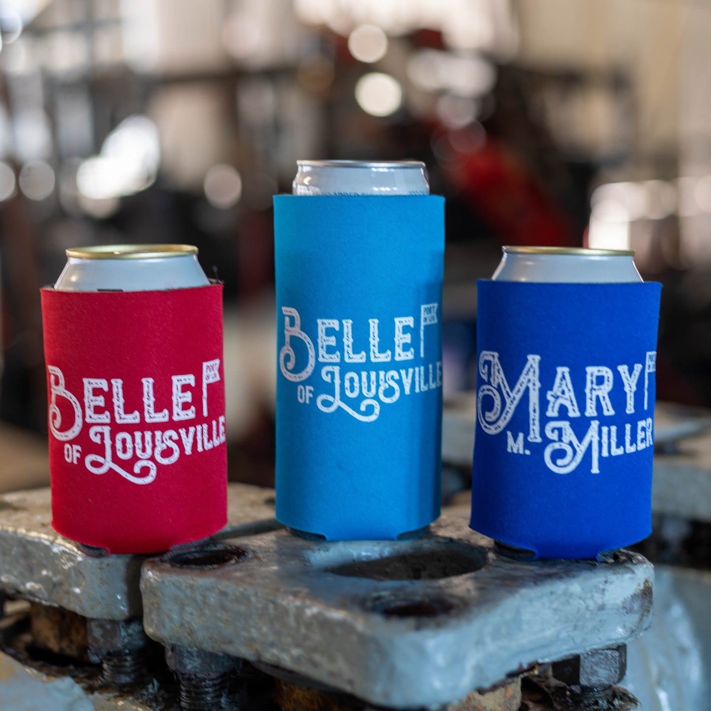 Three foam coozies on drink cans; left is red with white "Belle of Louisville" text on a regular 12oz can, middle is light blue with white "Belle of Louisville" text on a tall, slim 12oz can, right is dark blue with white "Mary M Miller" text on regular 12oz can
